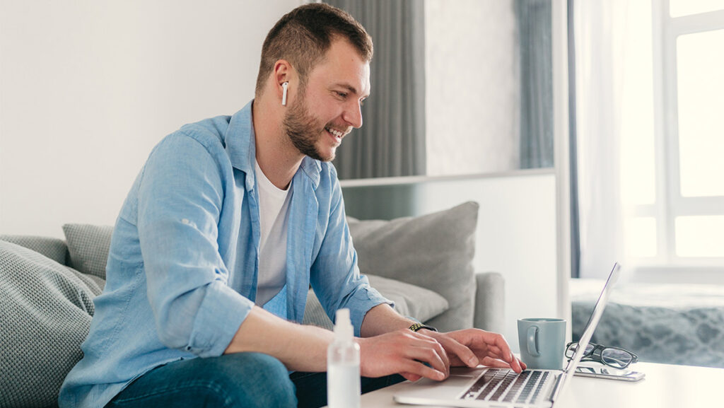 Hombre blanco con camisa azul sentado en el sofá de su casa trabajando en computadora
