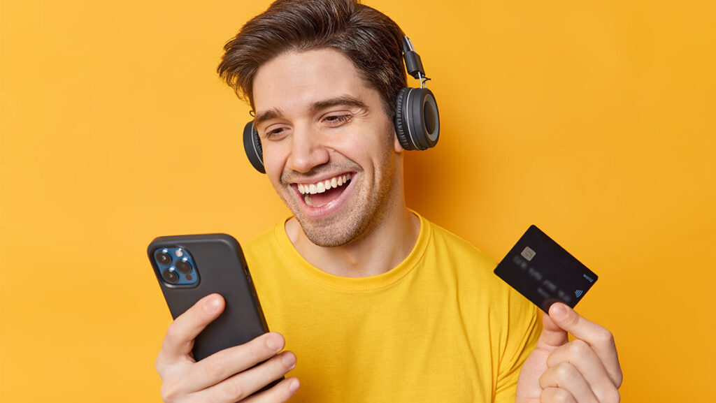 Joven sonriendo con audífonos negros y realizando compras o apuestas en línea a través de su smartphone