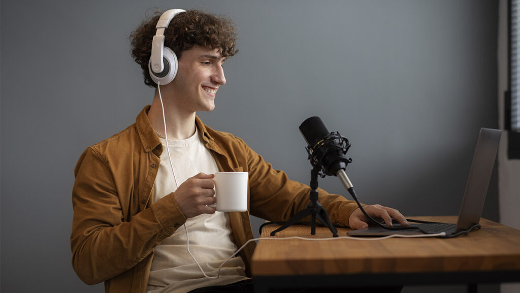 Hombre joven con audífonos, micrófono y taza de café frente a una computadora