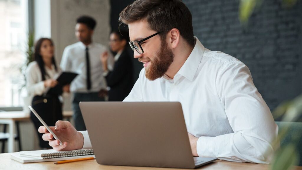 Hombre sonriente viendo su teléfono frente a computadora